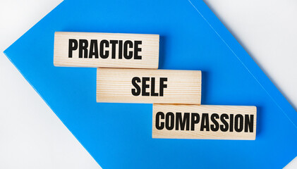 There is a blue notebook on a light gray background. Above are three wooden blocks with the words PRACTICE SELF COMPASSION