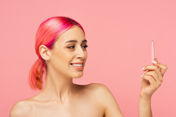 cheerful young woman with colorful hair looking at tweezers isolated on pink