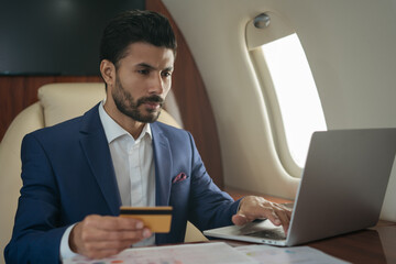 Handsome Arabian businessman holding credit card using laptop, shopping online sitting in airplane. Confident middle eastern entrepreneur flying luxury private jet. Successful business concept