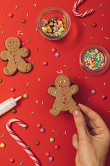 Christmas gingerbread cookie preparation process with gingerbread man, icing, sugar sprinkles and candy canes on a red table background. New year wallpaper flat lay.