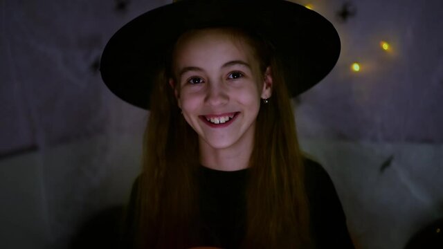 Children's halloween. A smiling girl in a witch's hat and a black T-shirt plays with an orange smiling balloon. Lights in the back dark background. Holiday at home.
