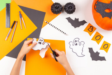 Preparing for Halloween. Teenage kid hands cutting colored paper with scissors and making Halloween decorations over white desk. Top view.