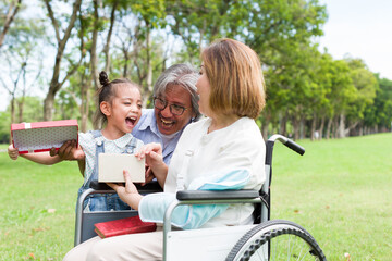 Cheerful Asian grandfather opening gift box with little girl while patient grandmother sitting on...