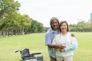 Asian elderly husband takes care his elderly sick wife while sitting on a wheelchair with injured arm wrapped in cast in the park. Asian senior couple spending time and relaxing together outdoor