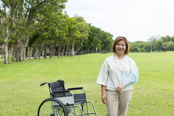 Asian patient elderly woman with injured arm wrapped in the cast standing near wheelchair in the park