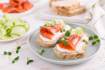 Toasts or open sandwiches with salted salmon, ricotta cream cheese, cucumber, sprouts and whole grain bread on white table. Healthy food, diet. Breakfast.