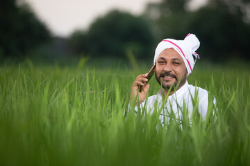 Portrait of happy young Indian man farmer sitting in agriculture land surrounded by crop holding...