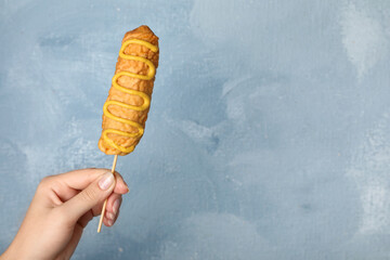 Woman holding delicious corn dog with mustard on light blue background, closeup. Space for text