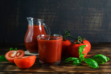 Tomato juice in a glass glass on dark wooden boards. A jug with tomato juice, basil leaves and sliced tomatoes in the background