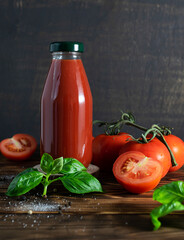 Tomato juice in a glass bottle on a dark wooden table. Whole and chopped tomatoes, basil leaves, salt and pepper peas in the background