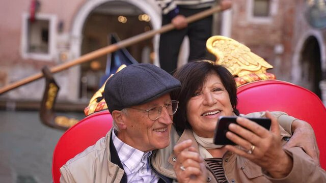 A cute romantic mature married couple rides on a gondola boat and takes a video call or selfies in Venice, in autumn fall, or winter. Happy holidays in Europe, Italy.