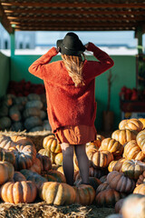 Back view of woman in brown sweater and hat on farmers market among stack of orange pumpkins. Cozy autumn vibes Halloween, Thanksgiving day