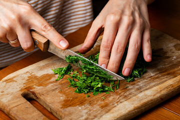 Woman cuts dill on a wooden cutting board with a knife.