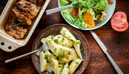 Boiled young potatoes with dill, fresh vegetable salad and baked chicken thighs on a dark wooden background.