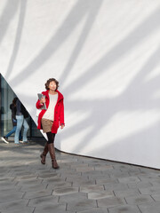 Curly woman in red duffle coat is walking by white wall. Smiling student with laptop. Freelancer with portable device for work. Modern lifestyle.