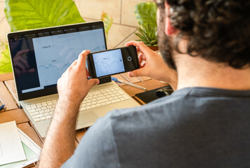 Young man using smartphone to take photo of charts on computer while sitting at table and investing in stock market at home office full of plants.