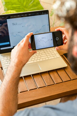 Young broker using smartphone to take photo of charts on computer while working remotely at home office full of plants.