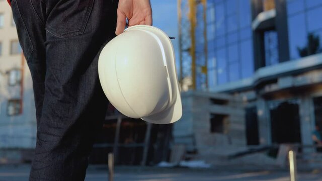 An Engineer-architect In A White Shirt And Orange Work Vest Stands Against The Backdrop Of A Modern Glass Building. Close-up View Of A Helmet In The Architect's Hand