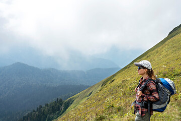 Fototapeta na wymiar Young woman hiker in cap and sunglasses with large hiking backpack looking at mountain view of the Aibga ridge of the Caucasus mountains, healthy active lifestyle, weekend activities beauty in nature