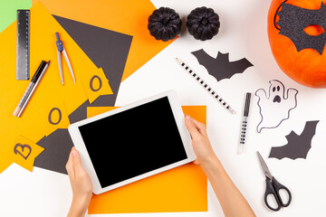 Preparing for Halloween. Teenage kid hands holding digital tablet computer. Colored paper, scissors, pumpkin, Halloween party decorations on white desk. Top view