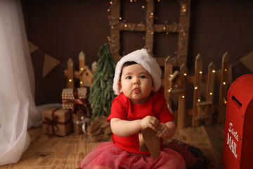 a cute dark-skinned girl is sitting on a wooden background and the background of a Christmas tree with gifts. a girl in a red suit, wearing a Santa hat, sends a letter to Santa Claus