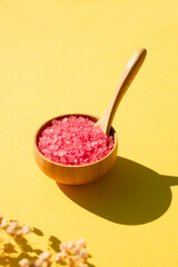 Wooden bowl with a spoon filled with pink bath sea salt. Beauty treatment for spa and wellness on yellow background.