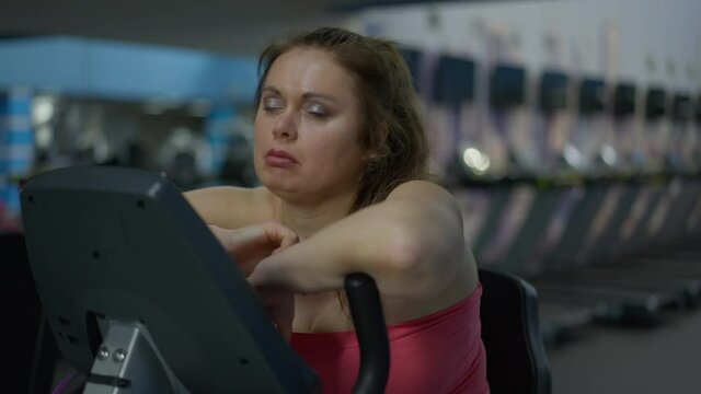 Sad tired overweight woman yawning sitting on exercise bike putting head on equipment. Portrait of exhausted plus-size Caucasian lady trying lose weight working out in gym indoors