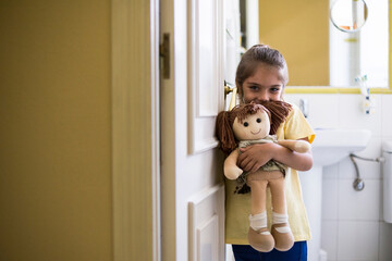 Portrait of smiling little girl standing in doorframe at home holding a doll