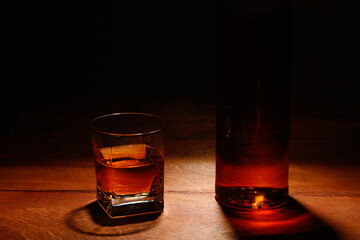 Glass of whiskey on a wooden table and part of the bottle on a dark background.