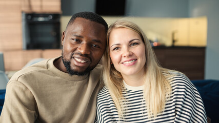 Portrait of loving diverse couple of Black man and Caucasian blonde woman looking at camera and smiling sitting on sofa at home. Loving biracial couple bonding and posing together