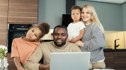 Portrait of beautiful loving diverse family of four looking at camera and smiling indoors. Black entrepreneur man working on laptop from home surrounded by White wife and two biracial kids