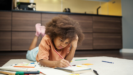 Creative biracial little girl lying on floor at home and drawing with pencils in notepad. Kid with...