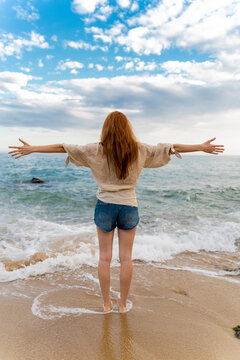 Back view of redheaded young woman standing at seafront with arms outstretched