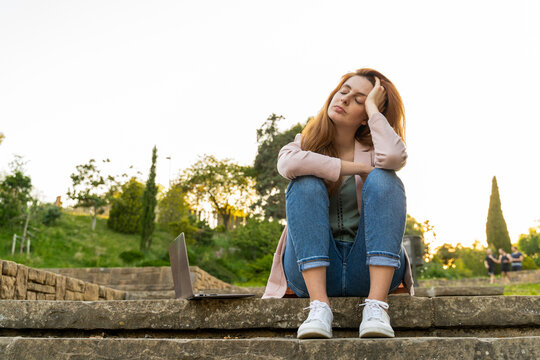 Young redheaded tired woman sitting on steps next to a laptop in a park