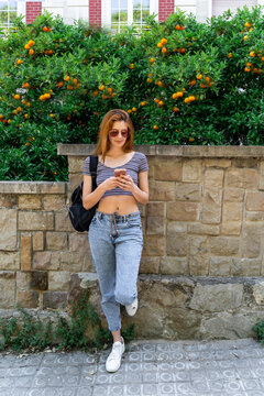 Woman with backpack leaning against stone wall using cell phone
