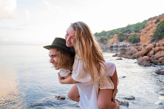 Young man giving his girlfriend a piggyback ride in front of the sea, Ibiza, Balearic Islands, Spain