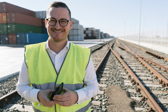 Portrait of smiling man on railway tracks in front of cargo containers with cell phone