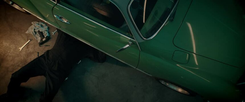 OVERHEAD High angle shot of Caucasian male mechanic repairing a vintage old car in a workshop, working under car bottom. Shot with 2x anamorphic lens