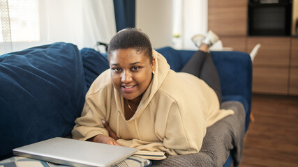 Portrait of smiling Black woman looking at camera lying on sofa at home with closed laptop nearby. Chubby female resting after working or studying remotely at self-isolation