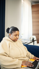 Side view of chubby Black woman in beige hoodie typing on laptop sitting on sofa at home. Focused female working or studying remotely using pc