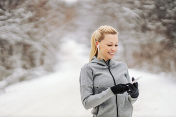Happy sportswoman standing in nature at snowy winter day and using her phone to search music. Technology, telecommunications, winter fitness