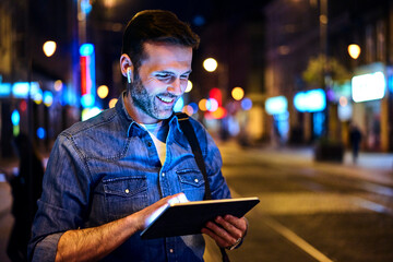 Smiling man with wireless headphones using digital tablet in the city at night
