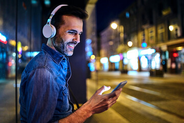 Smiling man with headphones using smartphone while waiting for night bus in the city