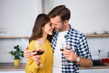 Happy couple standing in kitchen, with arms around, drinking red wine, kissing