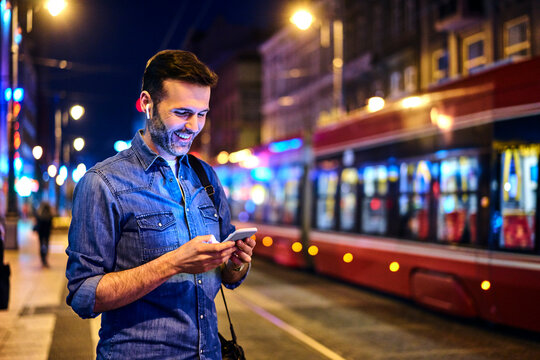 Smiling man with wireless headphones using smartphone while waiting for tram at night