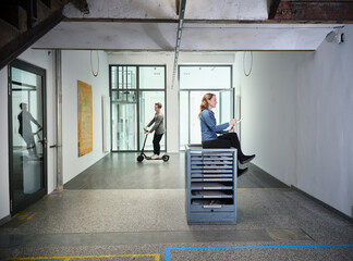 Businesswoman on office floor using tablet with colleague riding electric scooter in background