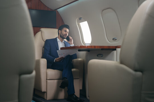 Handsome pensive middle eastern businessman talking on mobile phone, holding financial report working with documents sitting in plane. Confident entrepreneur flying private jet, successful business - Powered by Adobe