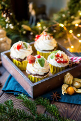 Cupcakes with cream and berries in a wooden box with a Christmas wreath and light bulbs. Cozy image with mucks for Christmas