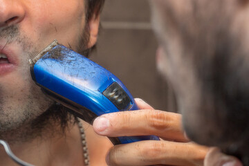 A man shaves his beard with an electric trimmer near the mirror