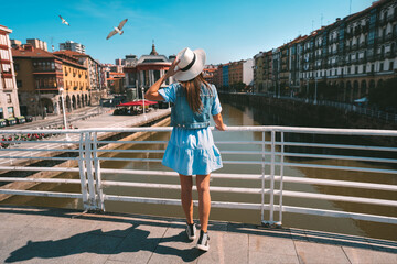 Rear view of young tourist woman in white sun hat walking in Bilbao city. Summer holiday vacation...
